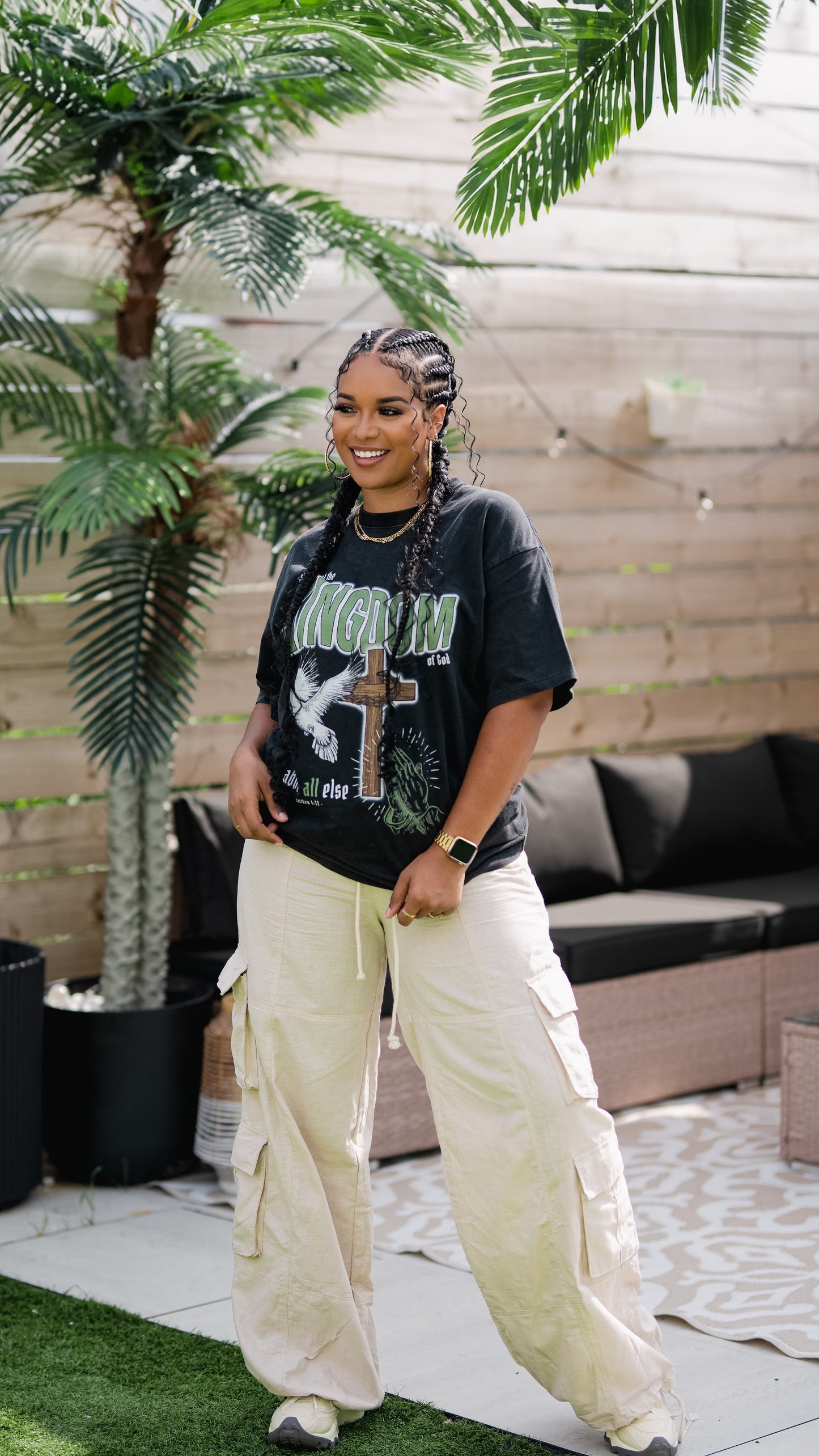 Person wearing a black graphic t-shirt and beige cargo pants standing outdoors with plants and a couch in the background.